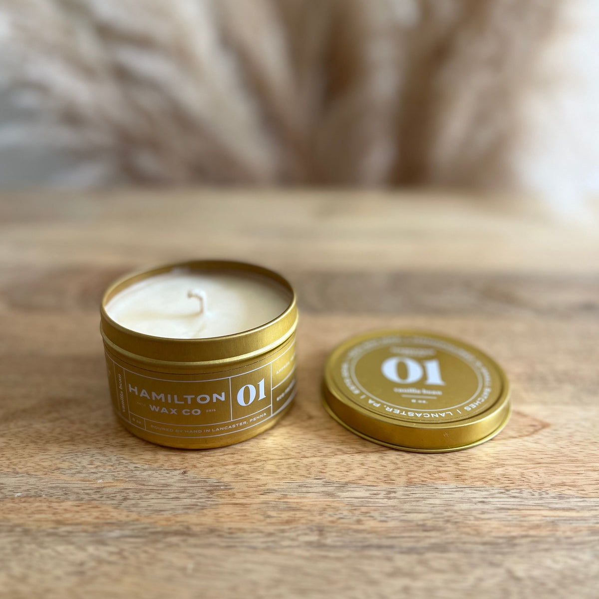 A small open gold metal candle tin with cream-colored soy candle sits on a wooden surface next to its matching lid. The label reads 'HAMILTON WAX CO' and '01'.
