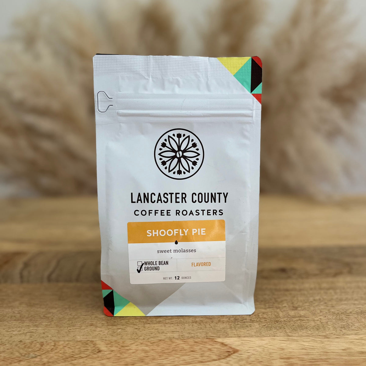 A white stand-up coffee bag labeled Lancaster County Coffee Roasters 'Shoofly Pie' ground coffee, standing on a wooden surface with a blurred beige background.