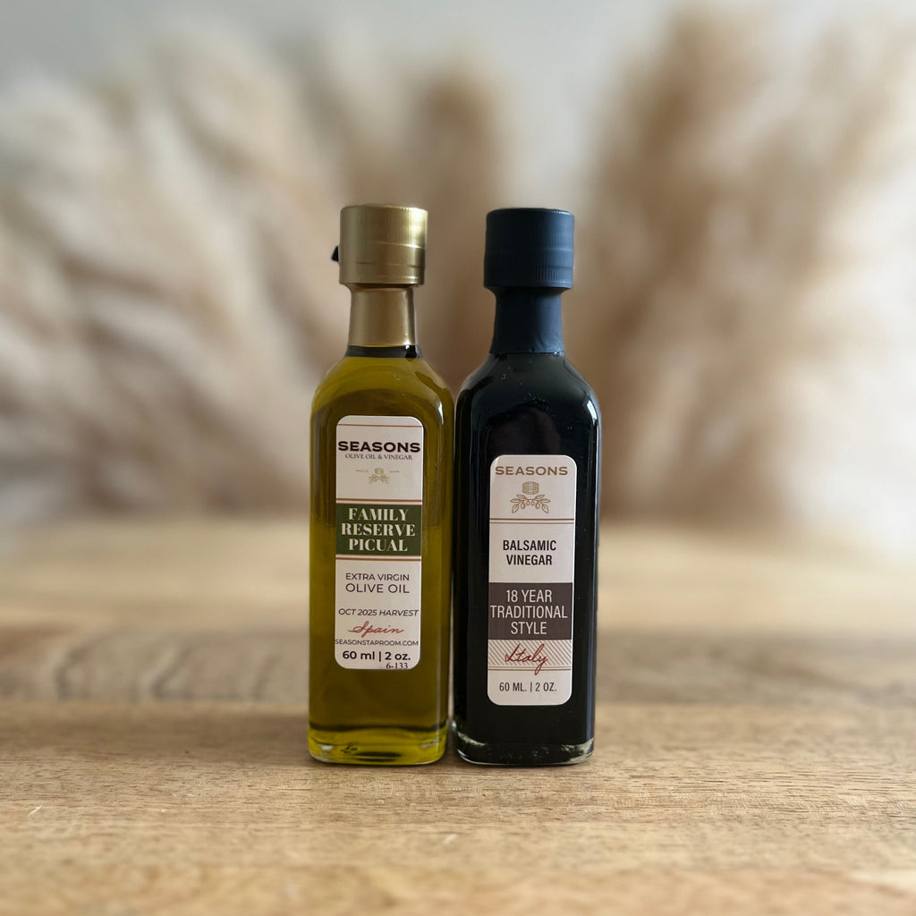 Two small countertop bottles of Seasons brand condiments standing on a wooden surface against a softly blurred neutral background with dried grasses. The left bottle is olive oil with a gold cap and green label, and the right bottle is dark balsamic vinegar with a black cap and dark label.