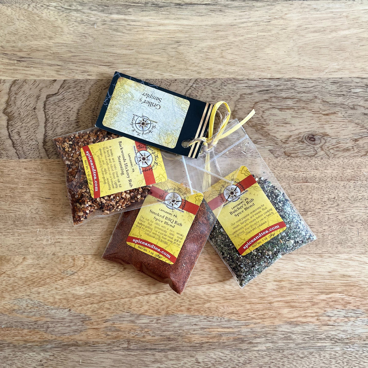 Three small clear plastic spice pouches with yellow and red labels are arranged on a light wood surface. One pouch is sealed with a black strip and yellow string, while the other two are open, showing reddish-brown and dark gray spice blends inside.