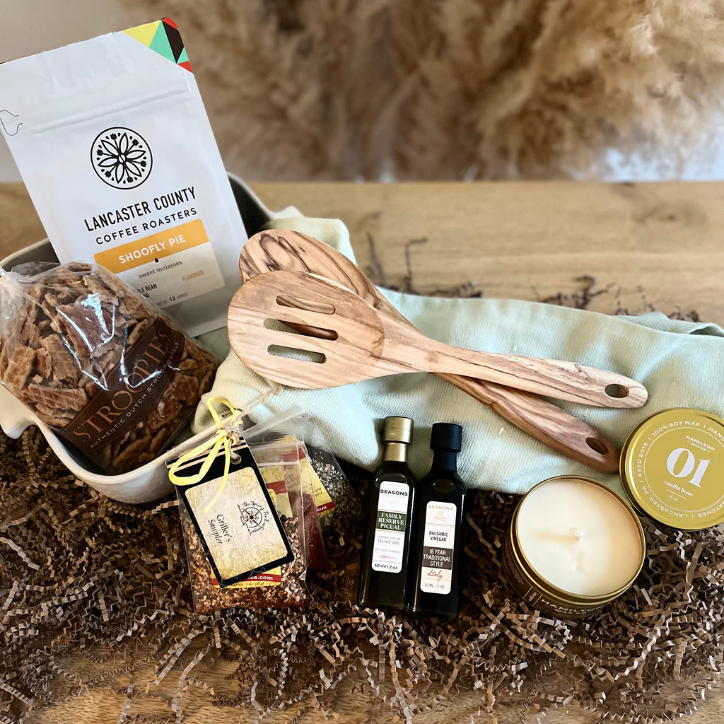 A curated gift basket arranged on a wooden surface with shredded paper filler. Visible items include a bag of Lancaster County Coffee Roasters coffee, two wooden utensils, a small candle, one olive oil bottle, one balsamic vinegar bottle, a package of stroopwaffle crumbles, and a round tin vanilla candle, with a pale green kitchen towel underneath.