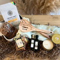 A curated gift basket arranged on a wooden surface with shredded paper filler. Visible items include a bag of Lancaster County Coffee Roasters coffee, two wooden utensils, a small candle, one olive oil bottle, one balsamic vinegar bottle, a package of stroopwaffle crumbles, and a round tin vanilla candle, with a pale green kitchen towel underneath.