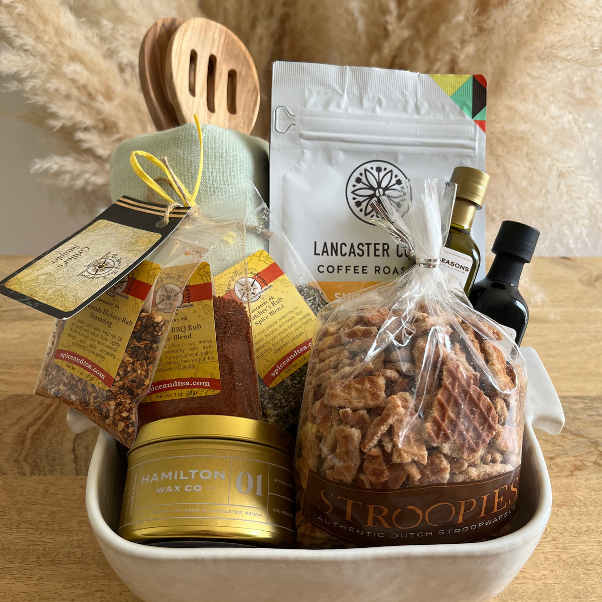 A curated gift basket in a white ceramic baking dish, filled with packaged food items including a large bag of Stroopies, a canister of Hamilton Wax Co. oil, Lancaster Coffee Roast coffee, and spice bags, styled with wooden spoons and pampas grass on a wooden table.