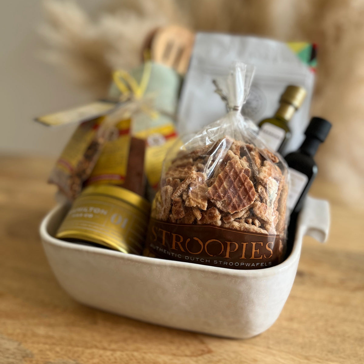 A gift basket arrangement in a white ceramic baking dish on a wooden surface, featuring a clear bag of Sroopies waffles, a gold tin candle, and small bottles, with a soft beige decorative background.