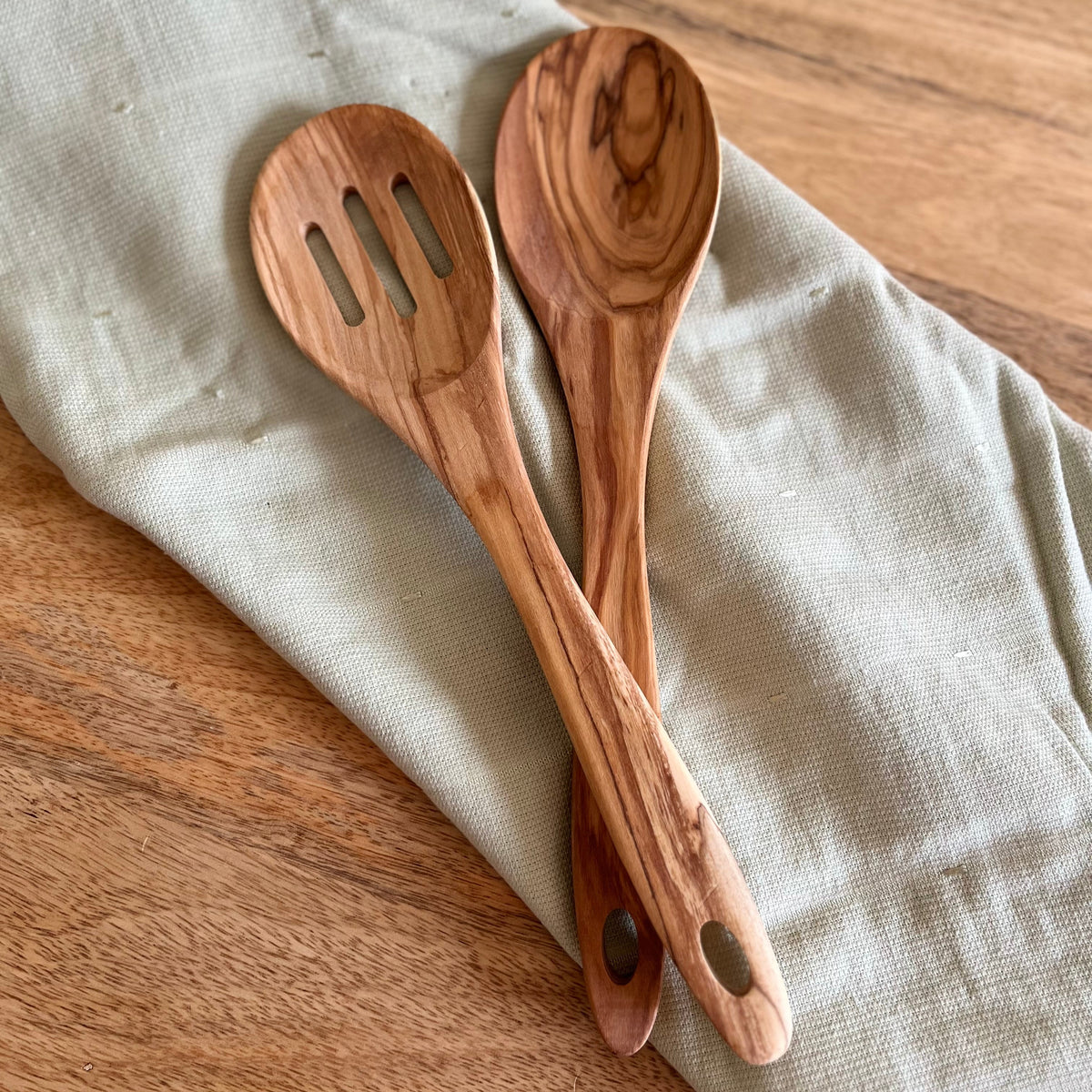 Two wooden kitchen utensils, a slotted spoon and a soup spoon, made from olive wood with distinctive grain patterns, resting on a light sage green linen cloth over a wooden table.